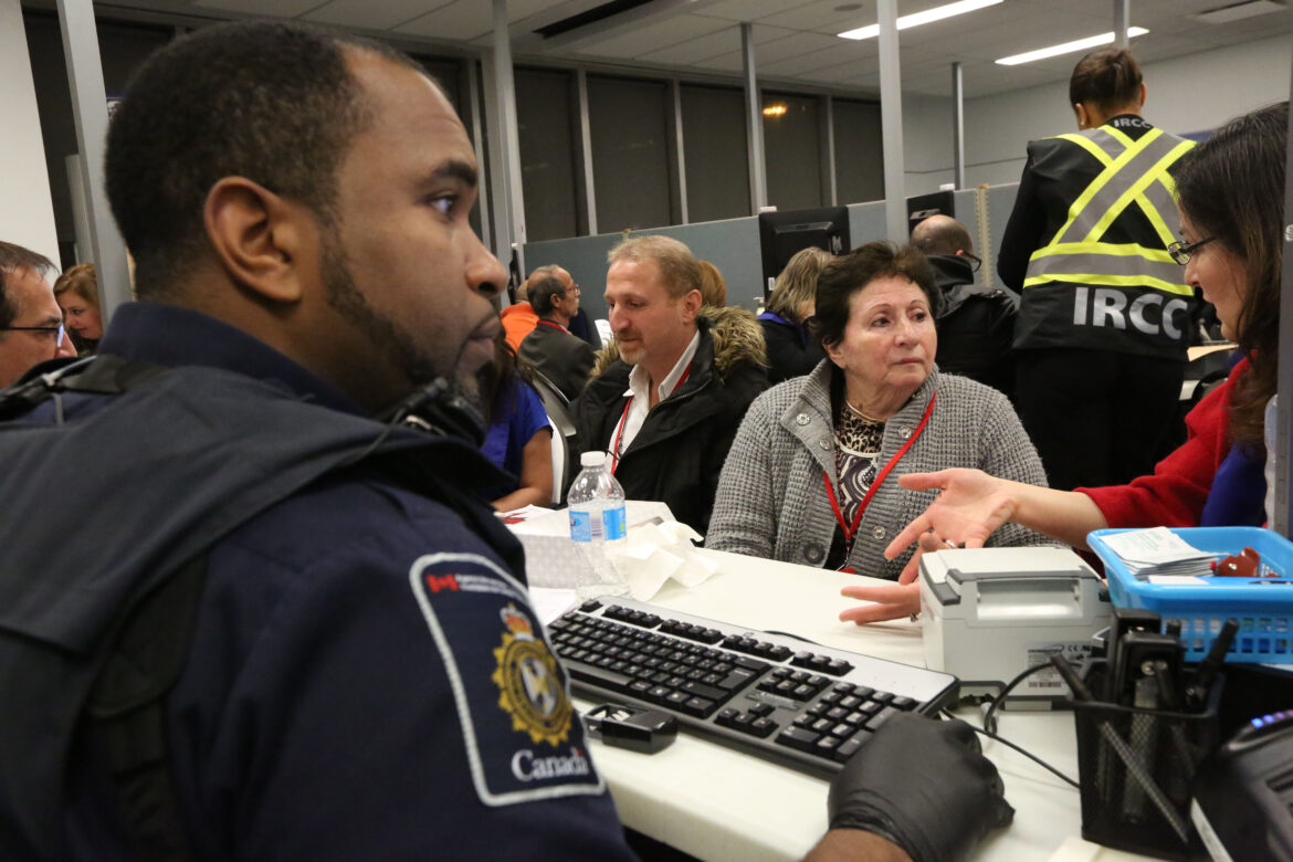 Border service desk scene: a uniformed officer with a Canada patch sits at a desk with a keyboard, talking to an elderly woman in a gray coat and red lanyard amid a busy service area.