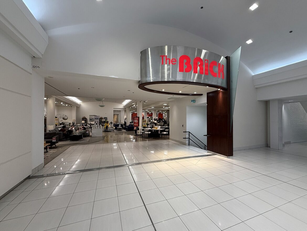 Wide mall corridor toward The Brick store with a curved metal sign; seating area visible on the left.