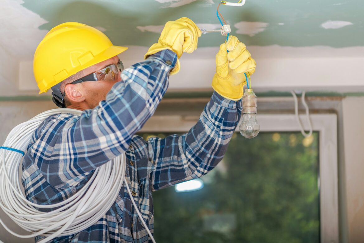 Electrician in a yellow hard hat and plaid shirt connects wiring to a light bulb fixture indoors, with a coil of cable over the shoulder.