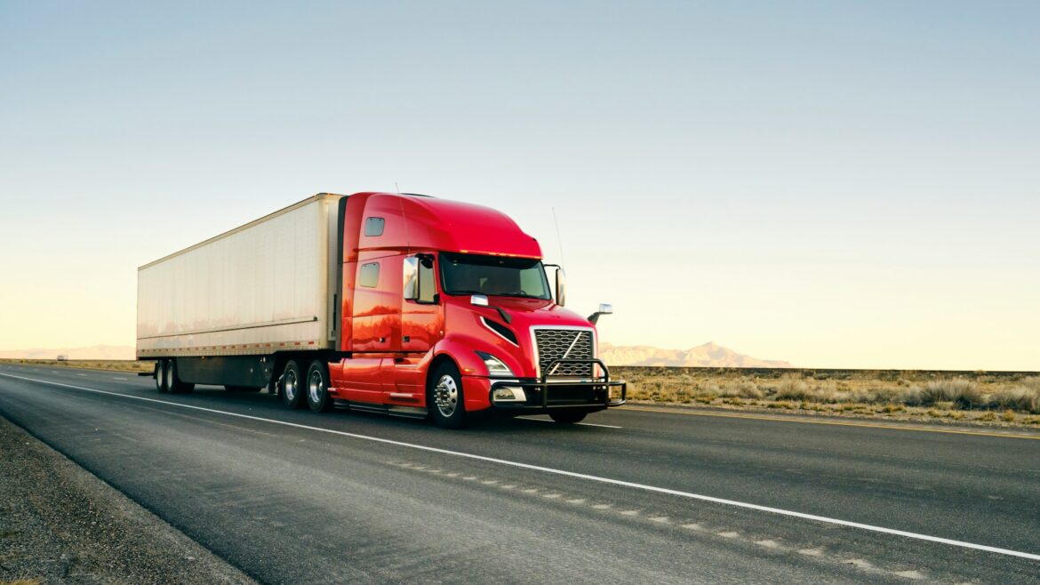 Red semi-truck with a white trailer driving on a straight desert highway under clear skies, mountains in the distance.