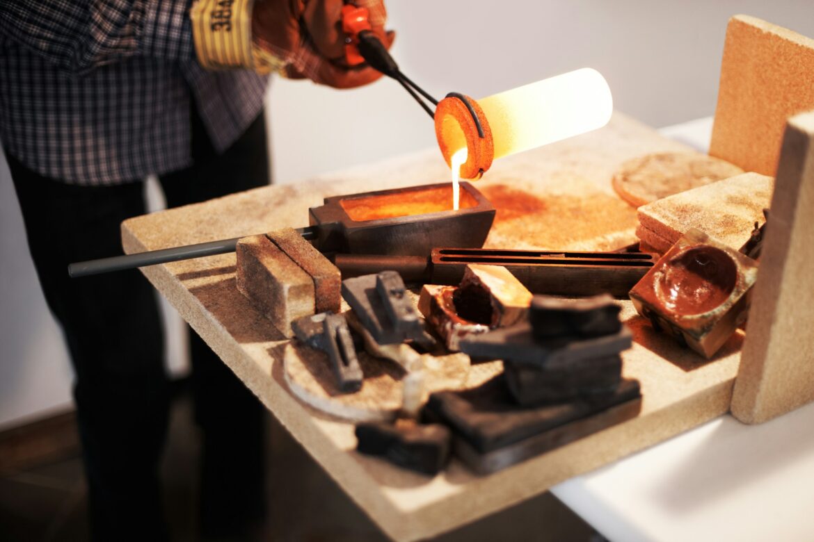 Person wearing gloves pours molten orange liquid from a crucible into a mold on a dusty workbench, with bricks and tools around.