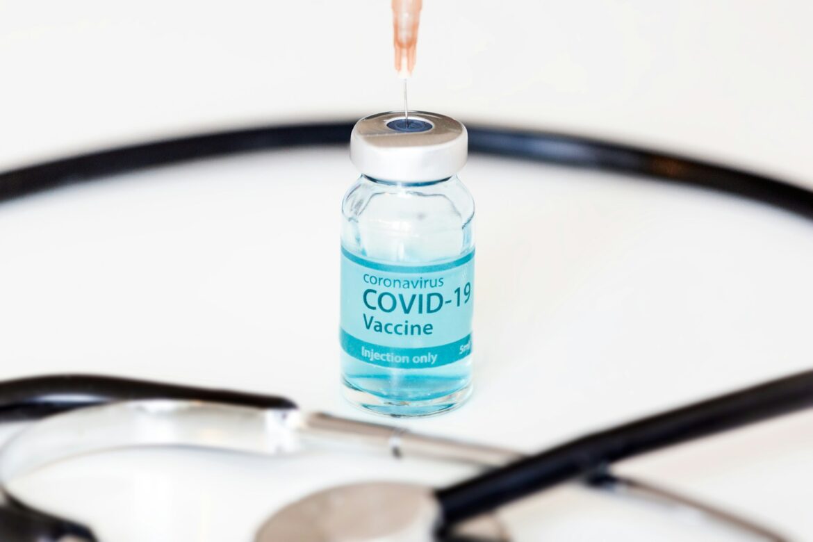 Blue-tinted vial labeled COVID-19 Vaccine with a syringe nearing the stopper, medical equipment blurred in the background