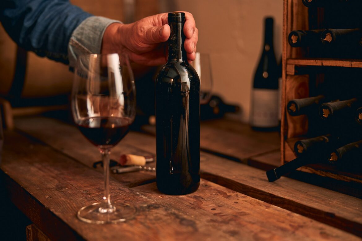 A dark wine bottle held by a hand over a rustic wooden table, with a glass of red wine nearby in a dim, cozy setting.