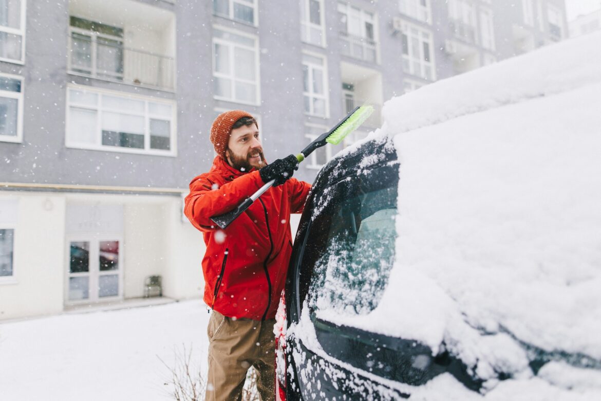 Man in an orange jacket and brown beanie brushing snow off a car windshield in a snowy urban scene.