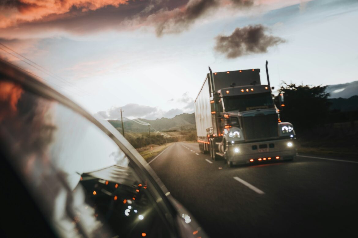 View from a car window showing a large silver semi-truck driving toward the camera on a two-lane highway at sunset.