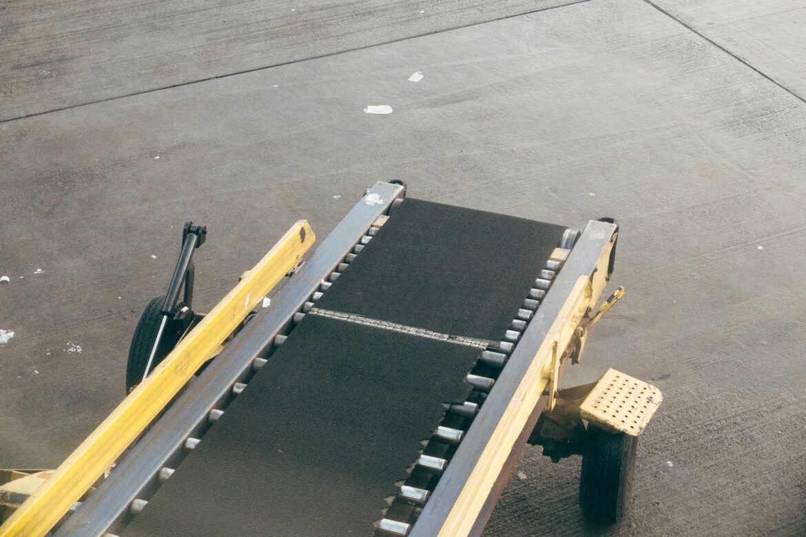 Portable conveyor belt on a yellow frame, extending over concrete ground, likely used for loading items onto a trailer