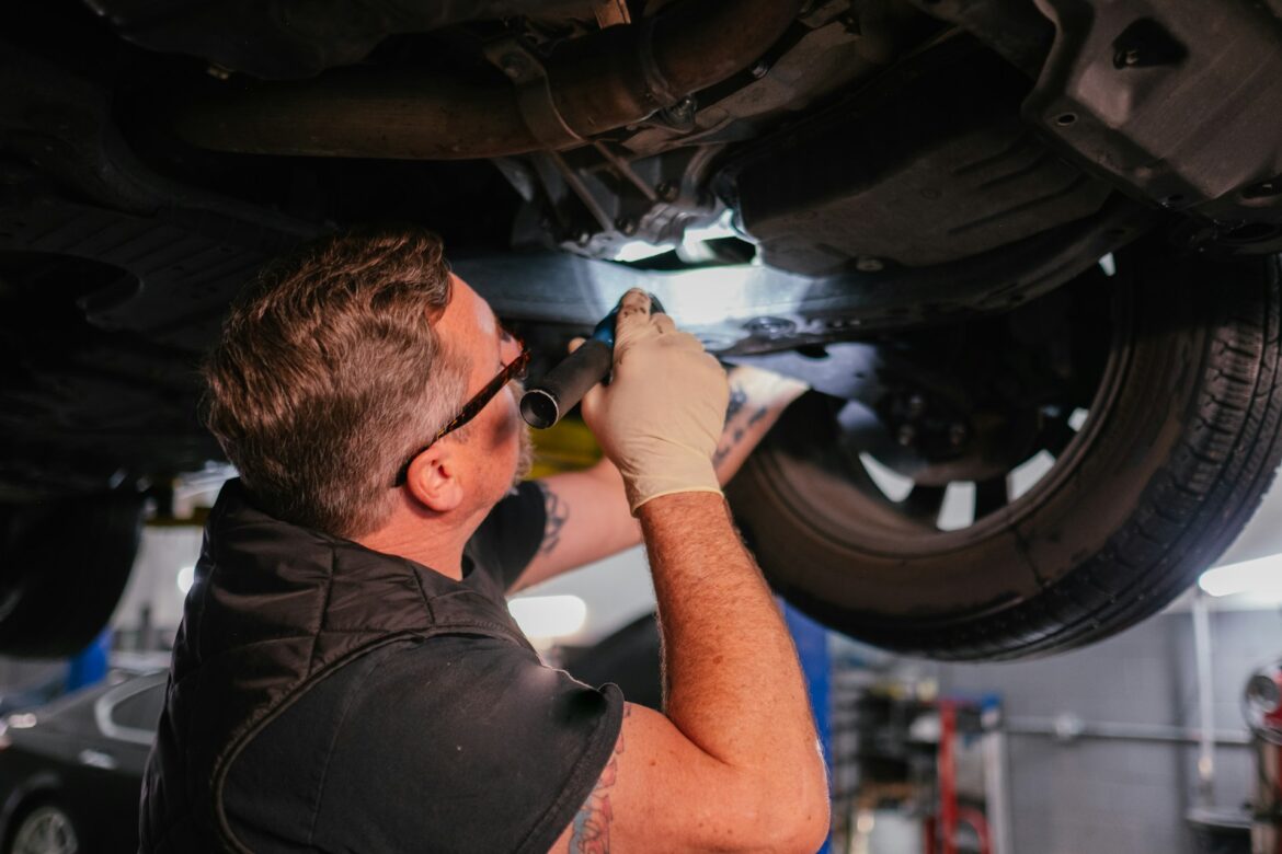 Mechanic in gloves under a raised car, using a tool to inspect the underside on a hydraulic lift.