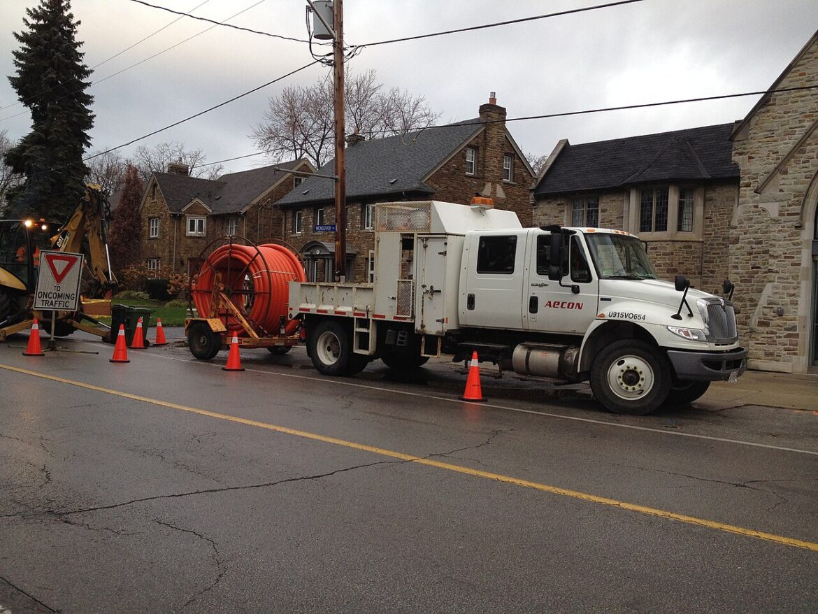 White utility truck with equipment bed and an orange cable reel on a trailer, parked on a wet street with traffic cones and a backhoe nearby in a residential, stone‑house neighborhood.