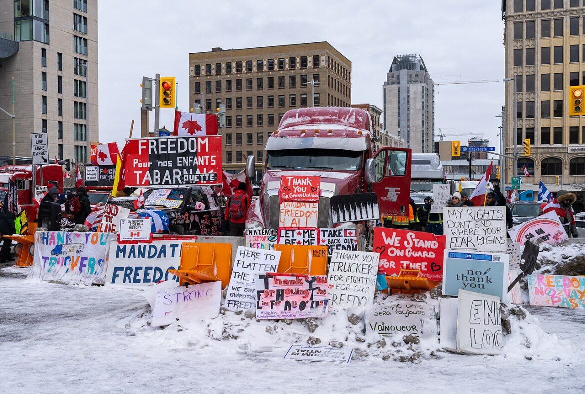 Snowy urban protest scene with a large red truck and protesters holding signs about freedom and mandates, Canadian flags visible behind barricades.