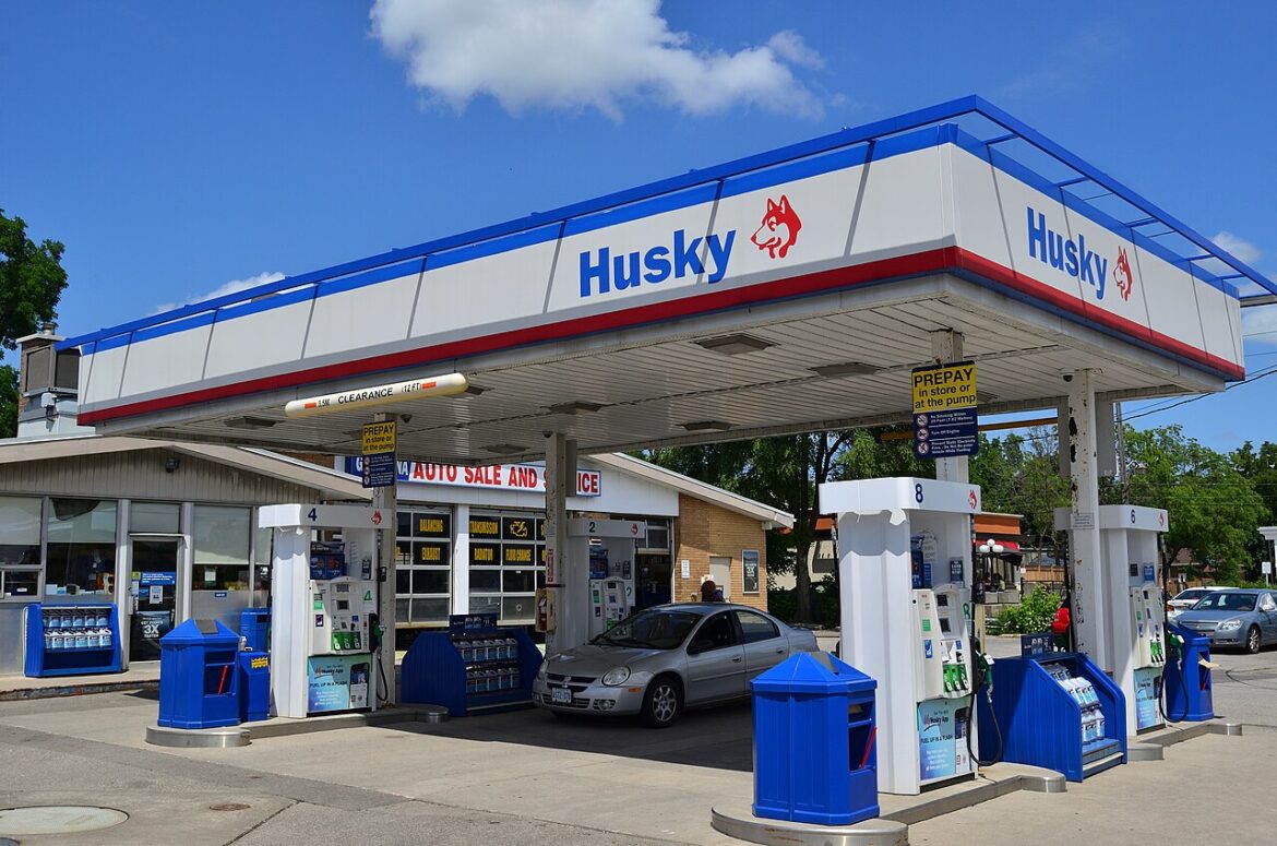 Gas station canopies with Husky branding and multiple fuel pumps; a silver car at one pump under a bright blue sky