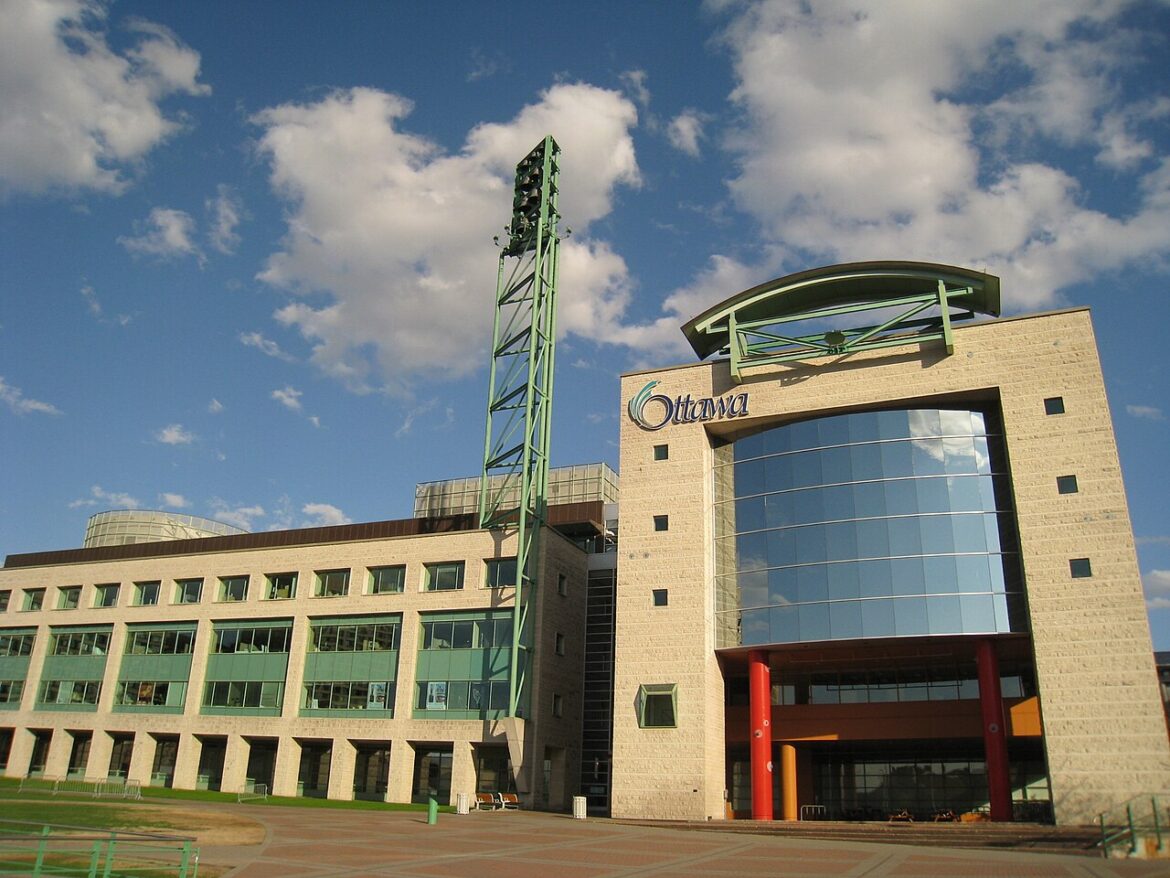 Modern brick building with a curved glass facade and a tall green lattice tower under a blue sky, Ottawa architectural complex.