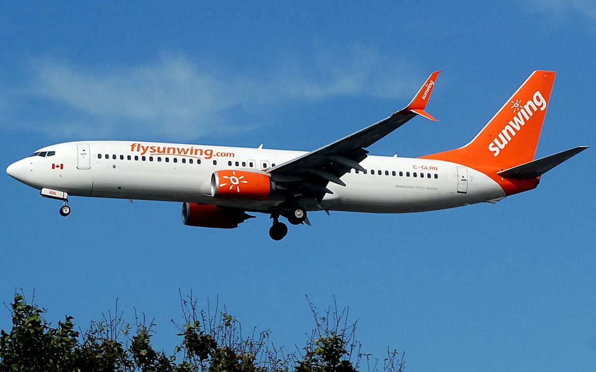 Sunwing Airlines jet in flight with orange tail and logo, gear down against a blue sky.