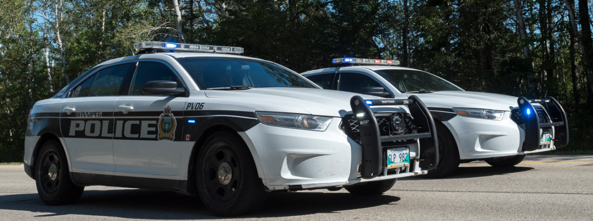 Two white police cars with rooftop lights parked on a road, front ends facing right, bold 'POLICE' lettering on the side and push bars on the front. Hint of a wooded area behind them.