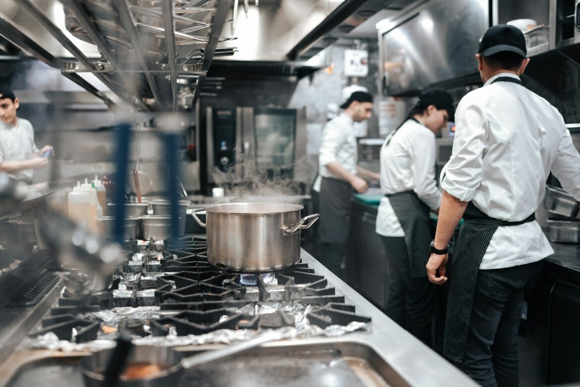Busy commercial kitchen with chefs in white jackets and aprons working around a large gas stove; steam rises from a simmering pot in the foreground.