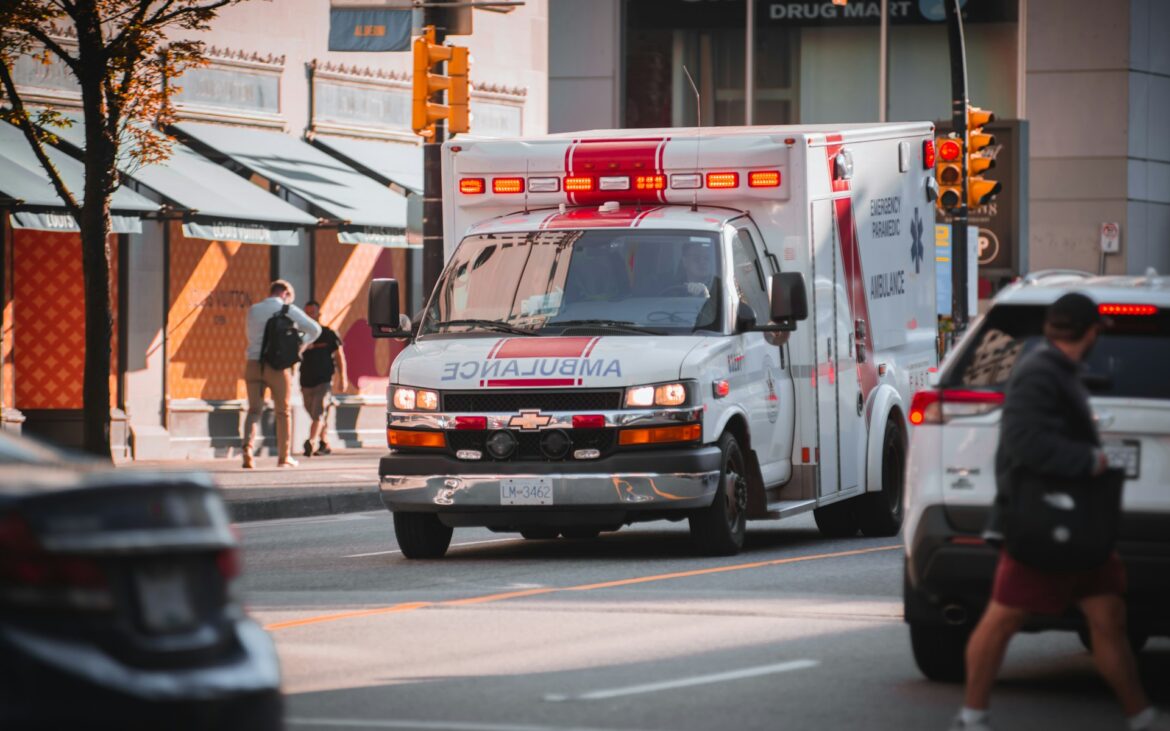Front view of a white ambulance with red flashing lights driving through a busy city street, pedestrians nearby and storefronts in the background.