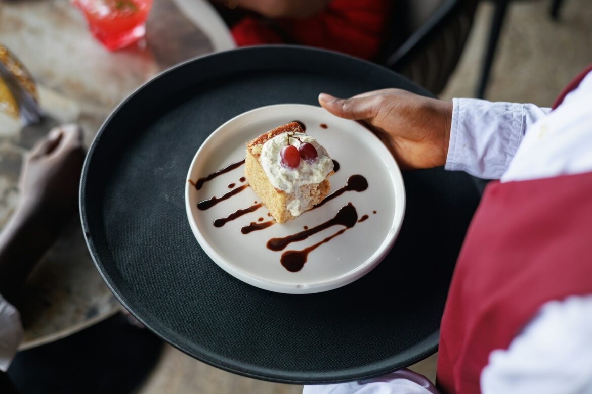 Slice of sponge cake topped with whipped cream and two red grapes on a white plate, drizzled with chocolate sauce, held by a server in a white shirt.