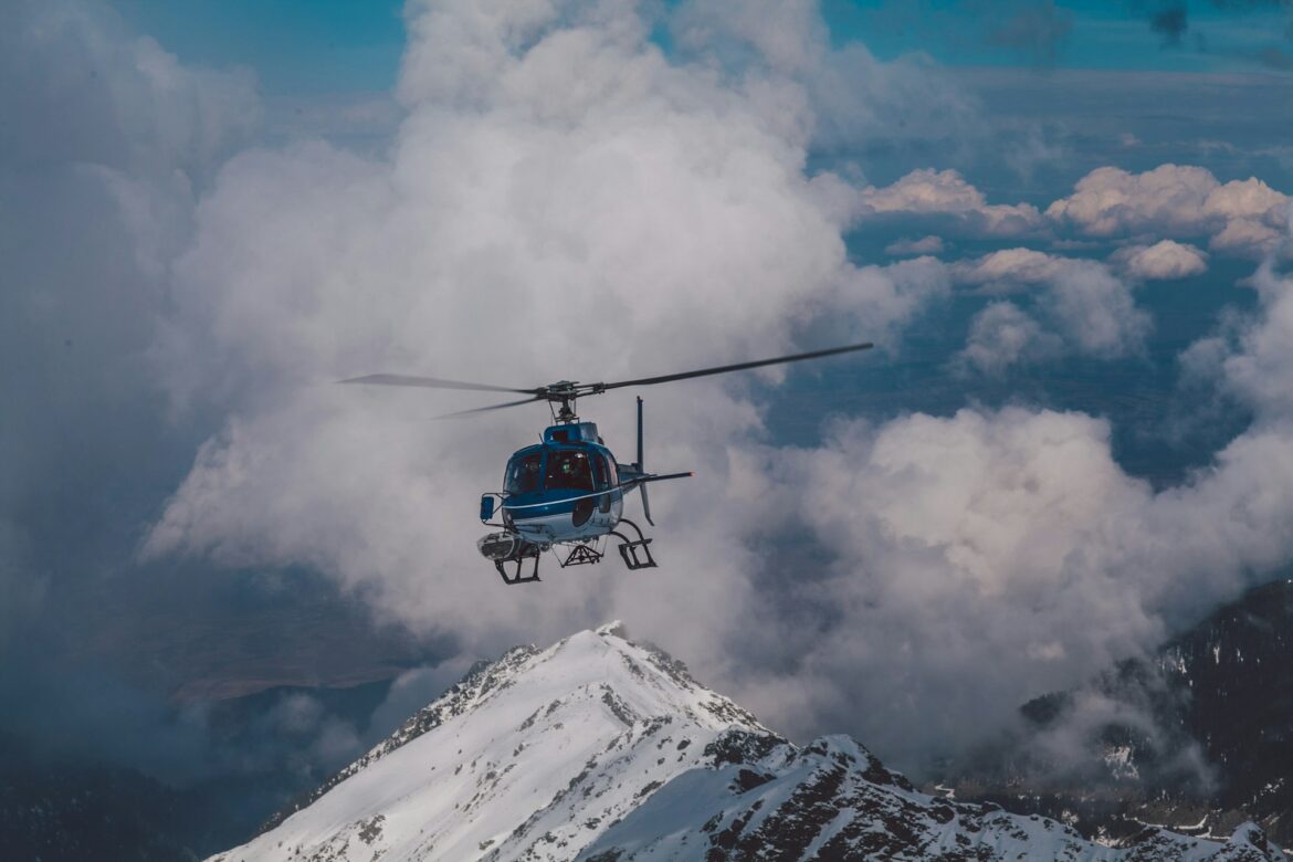 Blue helicopter flying among white clouds over snow-covered mountains.