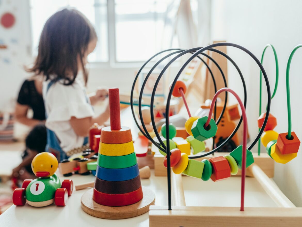 Close-up of a colorful bead maze and wooden stacker on a table, with a child playing in the softly blurred background.