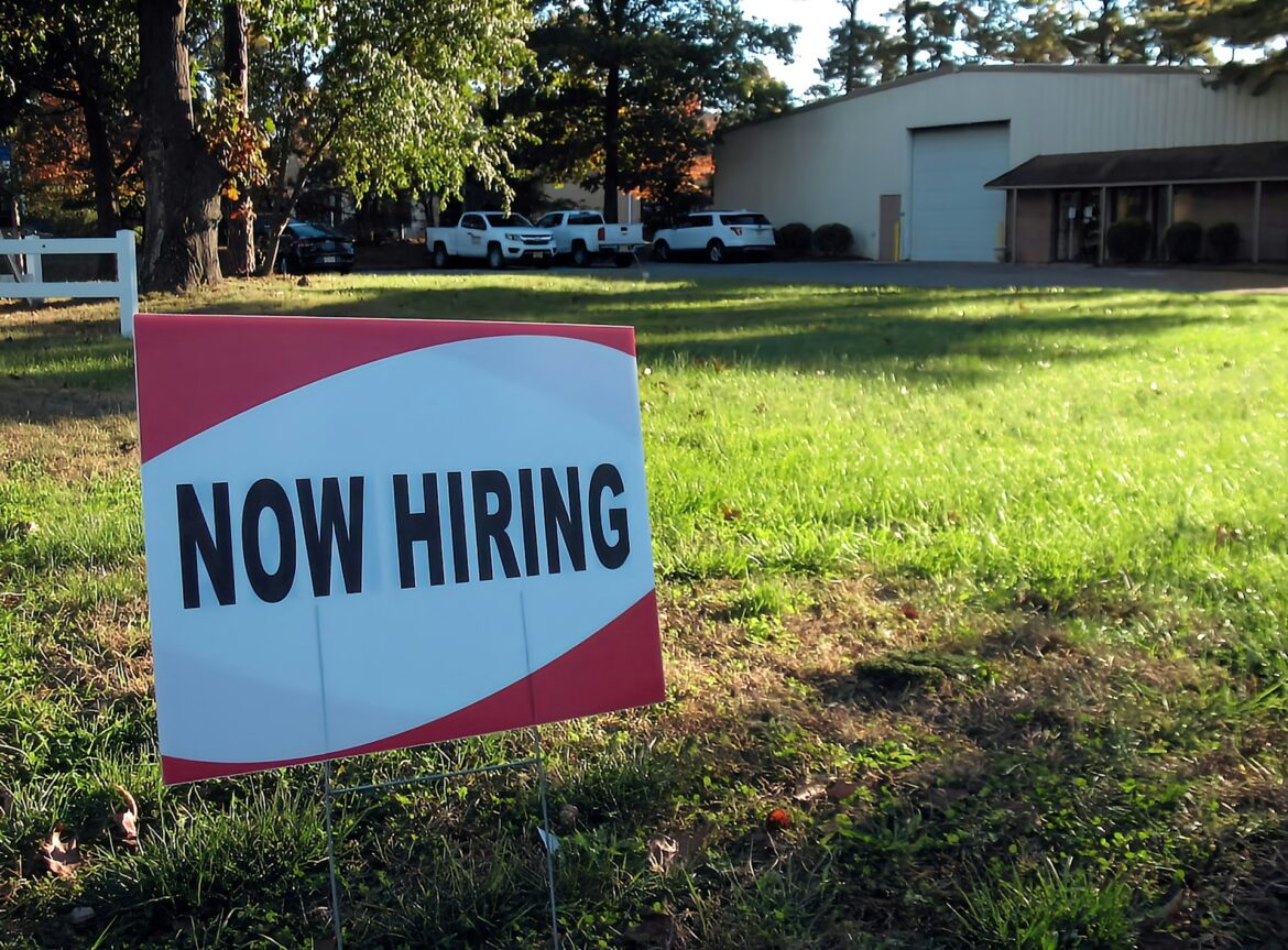 Now hiring sign in a grassy lot with an industrial building and parked vehicles in the background.