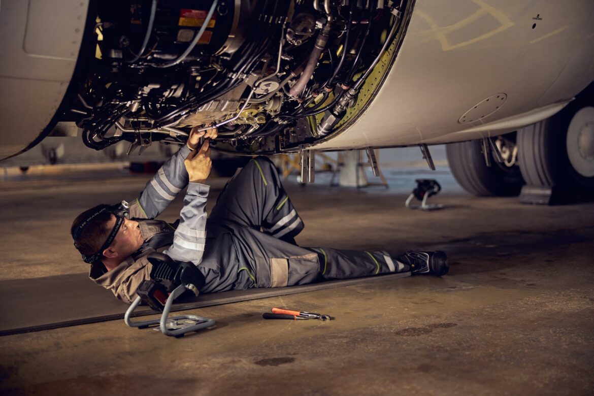 A mechanic in protective gear lying on the hangar floor under an aircraft engine, working with exposed wires and components.