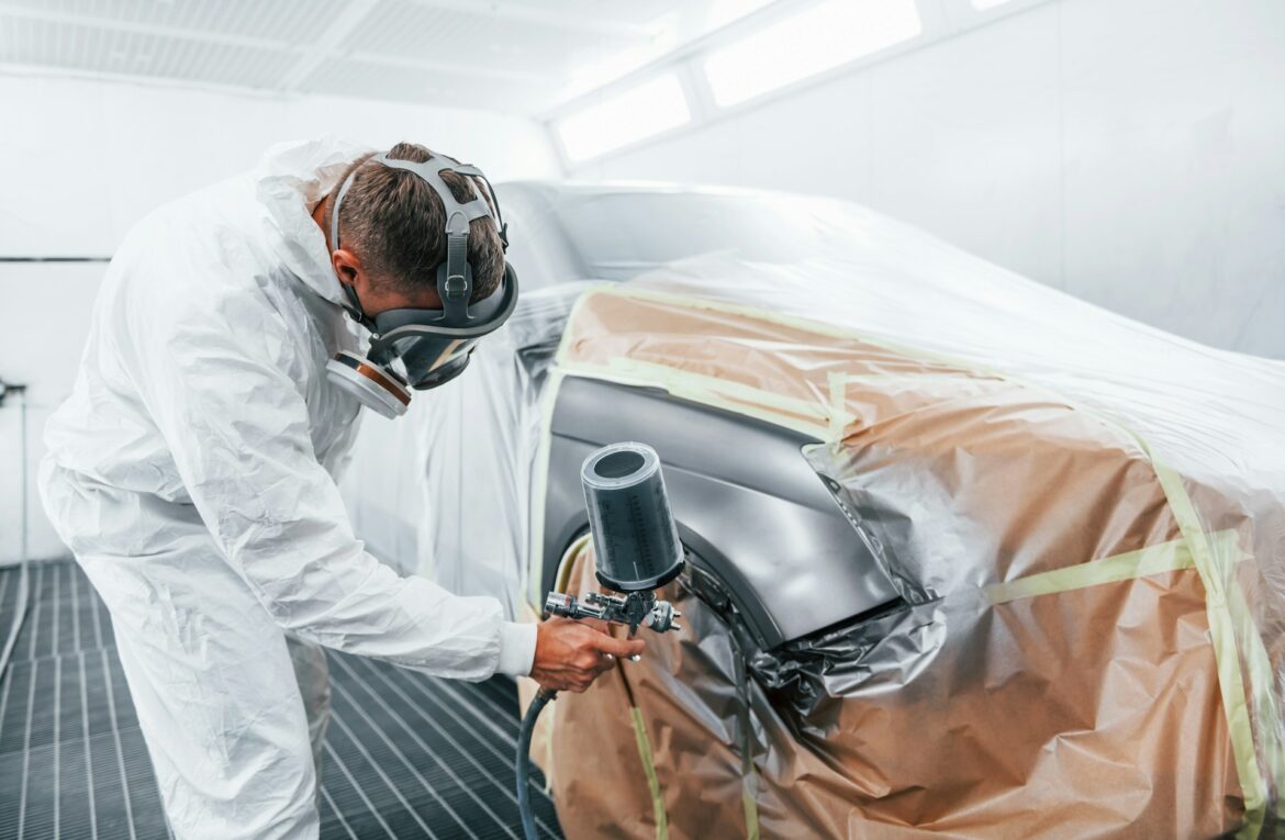 Worker in white protective suit and respirator hoods car in a spray booth, applying paint with a spray gun masking taped brown panels nearby