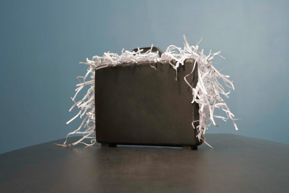 Black leather briefcase overflowing with white shredded paper on a dark table against a blue wall, suggesting discarded documents or clutter.