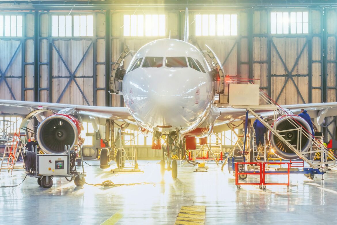 Front view of a white commercial airliner in a maintenance hangar with open service doors and crew at work.