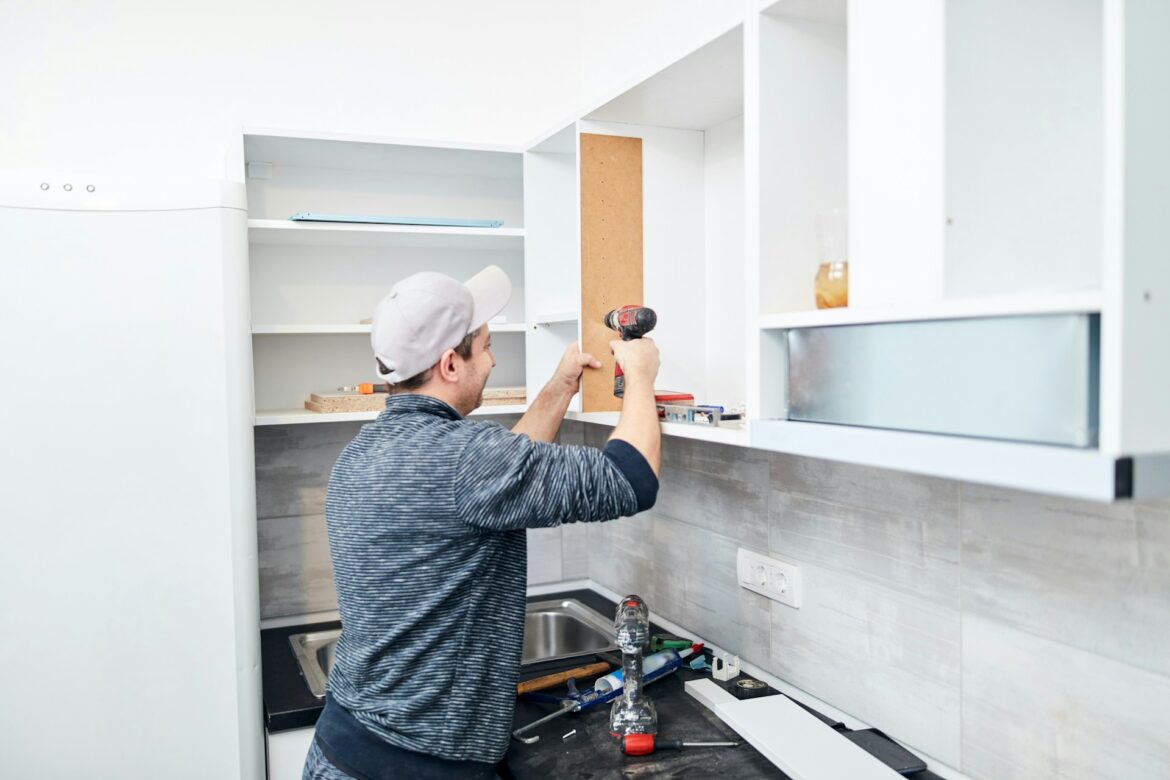 Man in a striped shirt installs a vertical cabinet panel with a drill in a modern kitchen.