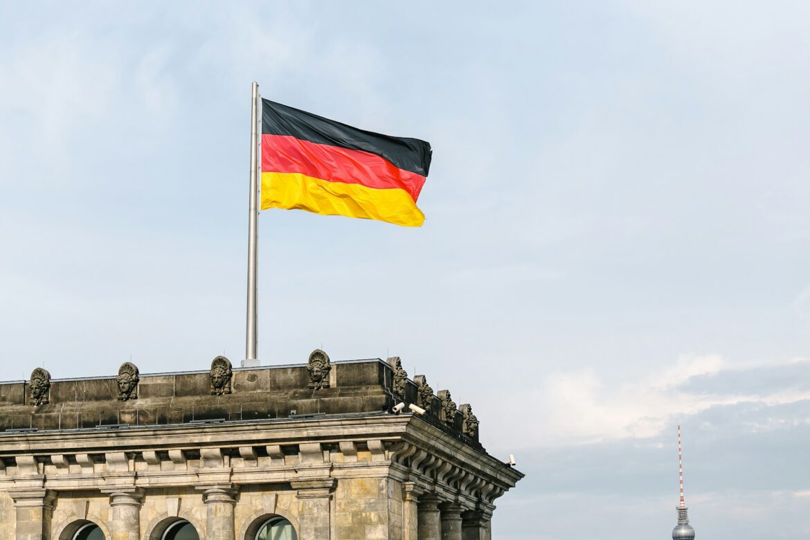 German flag waving atop a flagpole above a historic stone building with arches against a blue sky