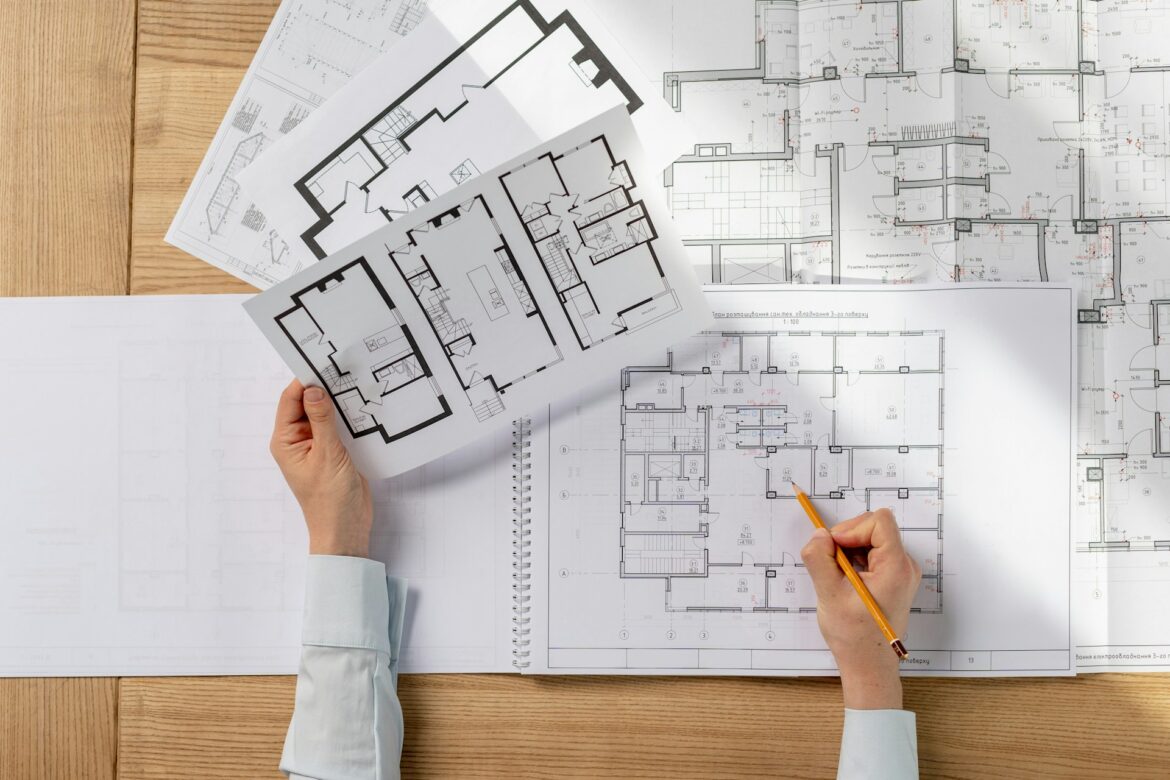 Architectural floor plans being reviewed, with a hand holding a sheet over a wooden desk and a pencil drawing on the plan.