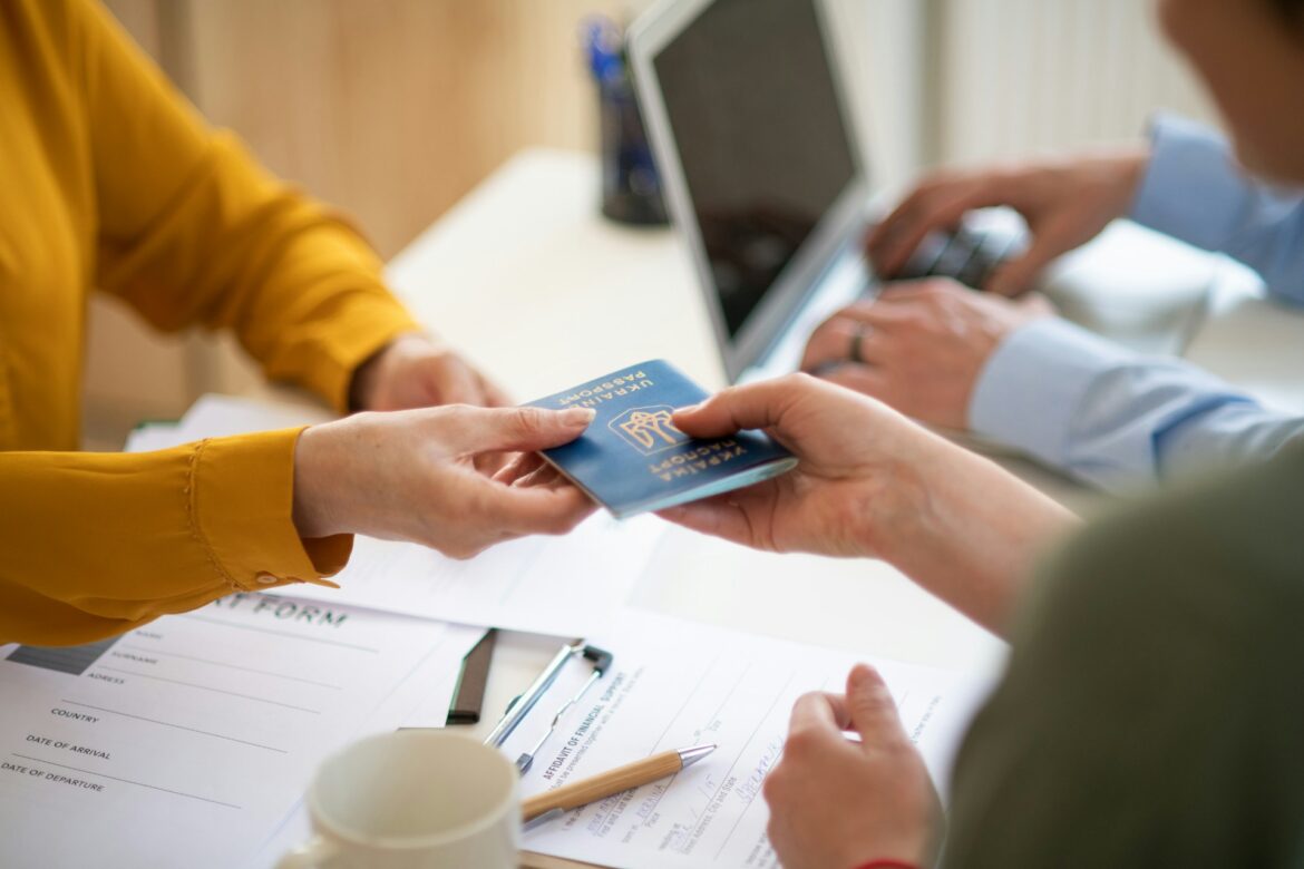 A blue passport is being handed from one person to another over a desk with forms and a mug nearby.