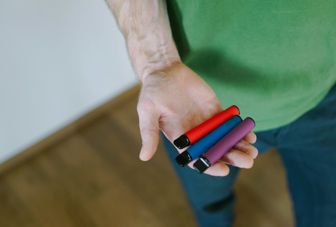A hand holds four colored markers (red, blue, purple, pink) with black caps visible against a green shirt.