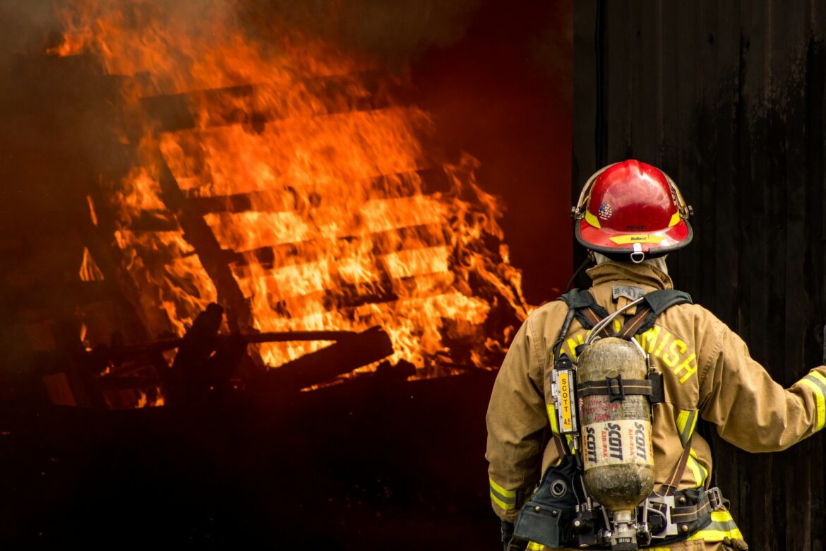 Firefighter in full turnout gear with helmet and breathing apparatus facing a blazing wall of flames and smoke.