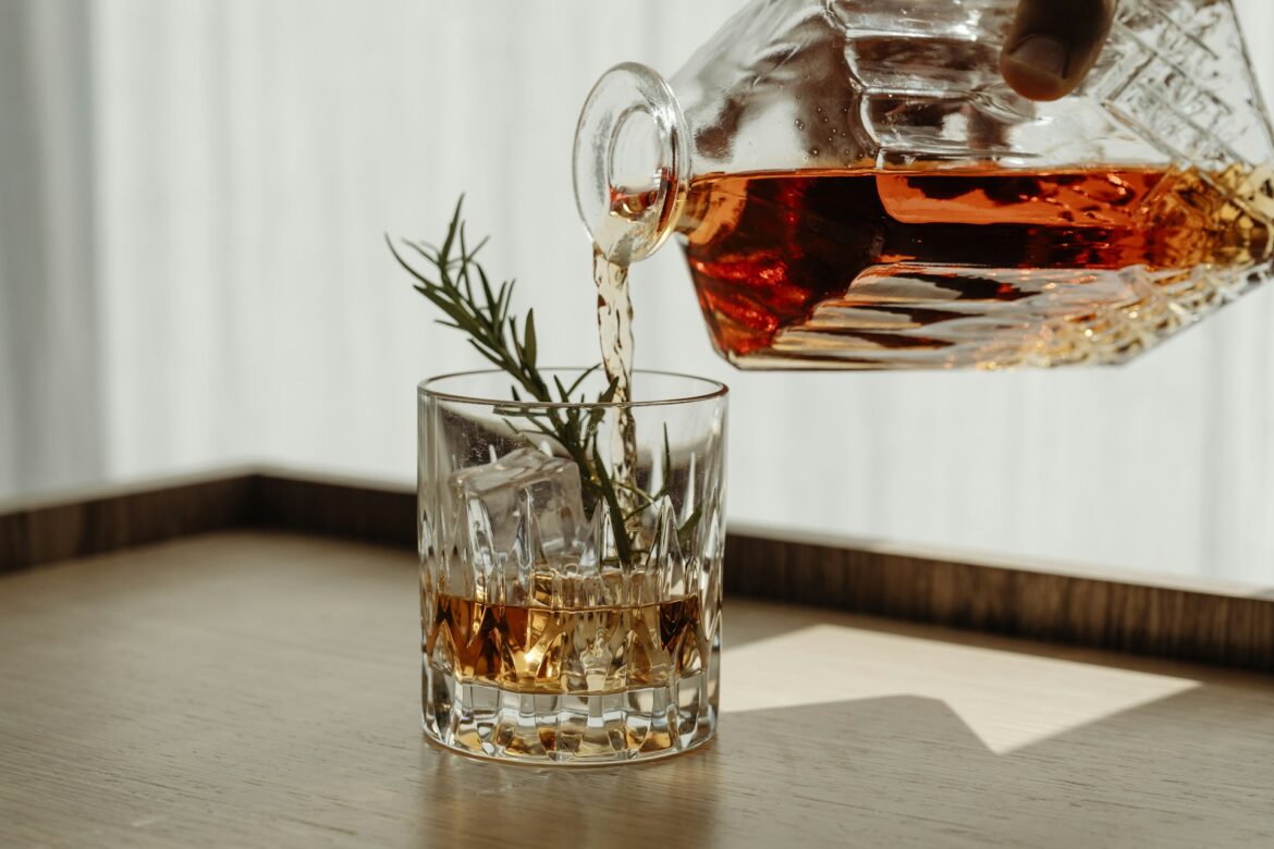 Pouring amber whiskey into a glass with ice and a rosemary sprig nearby on a wooden surface laid out for a drink.