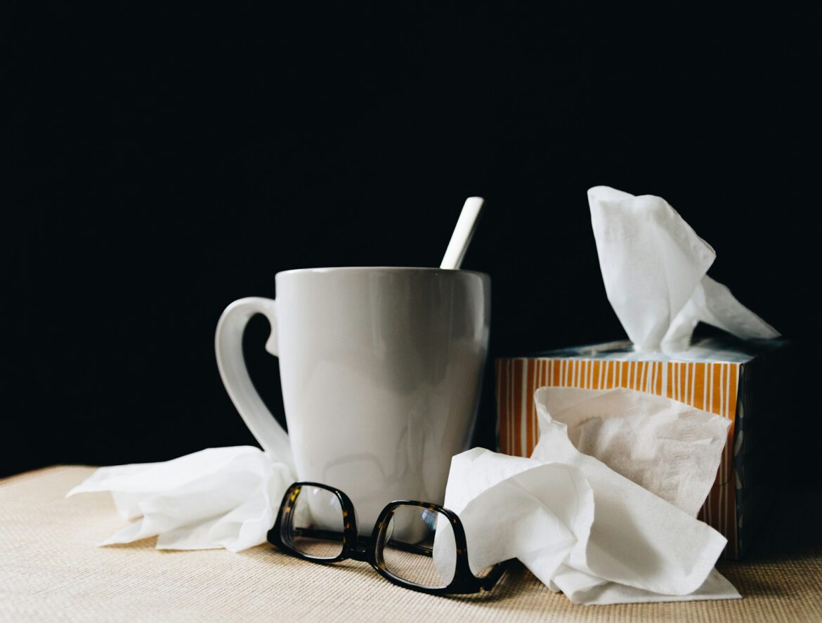 Messy desk scene with a white mug, tissue box, crumpled tissues, and black-framed glasses on a beige surface against a dark background.