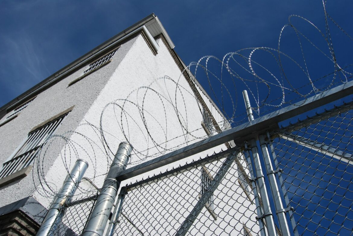 Barbed razor wire atop a chain-link fence around a white industrial building against a clear blue sky.