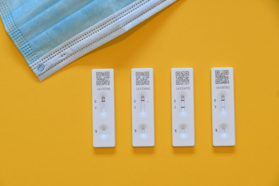 Blue disposable face mask folded at the top left next to four white COVID-19 rapid test cassettes on a yellow background.