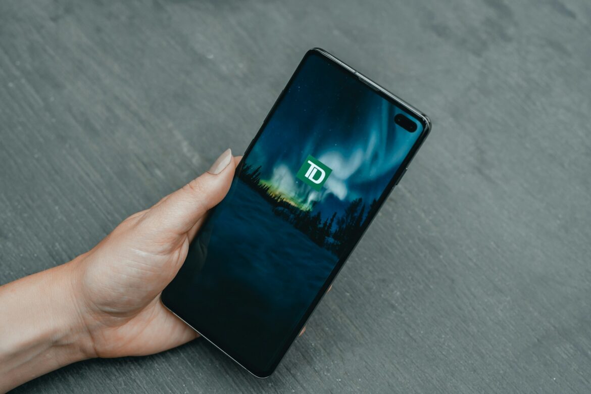 Close-up of a hand holding a smartphone showing a blue wallpaper with a green TD logo on the screen