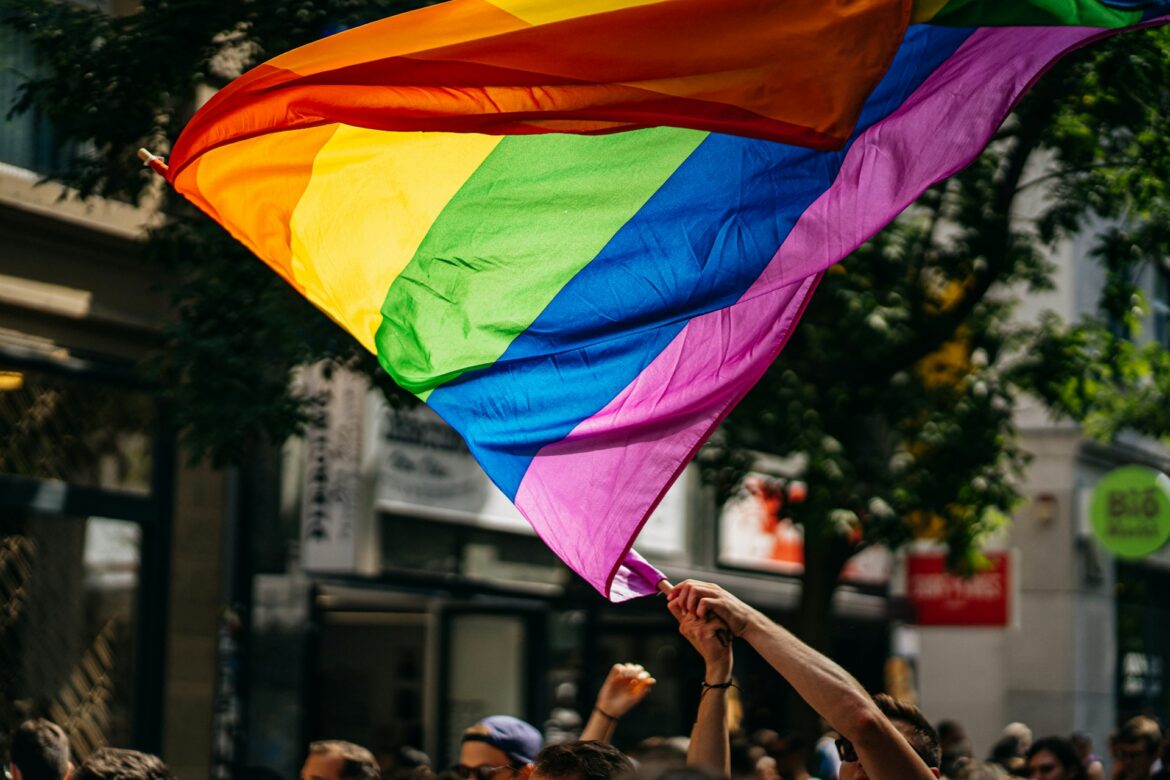 Pride flag being waved in a crowd during a street parade, hands raised beneath the colorful banner