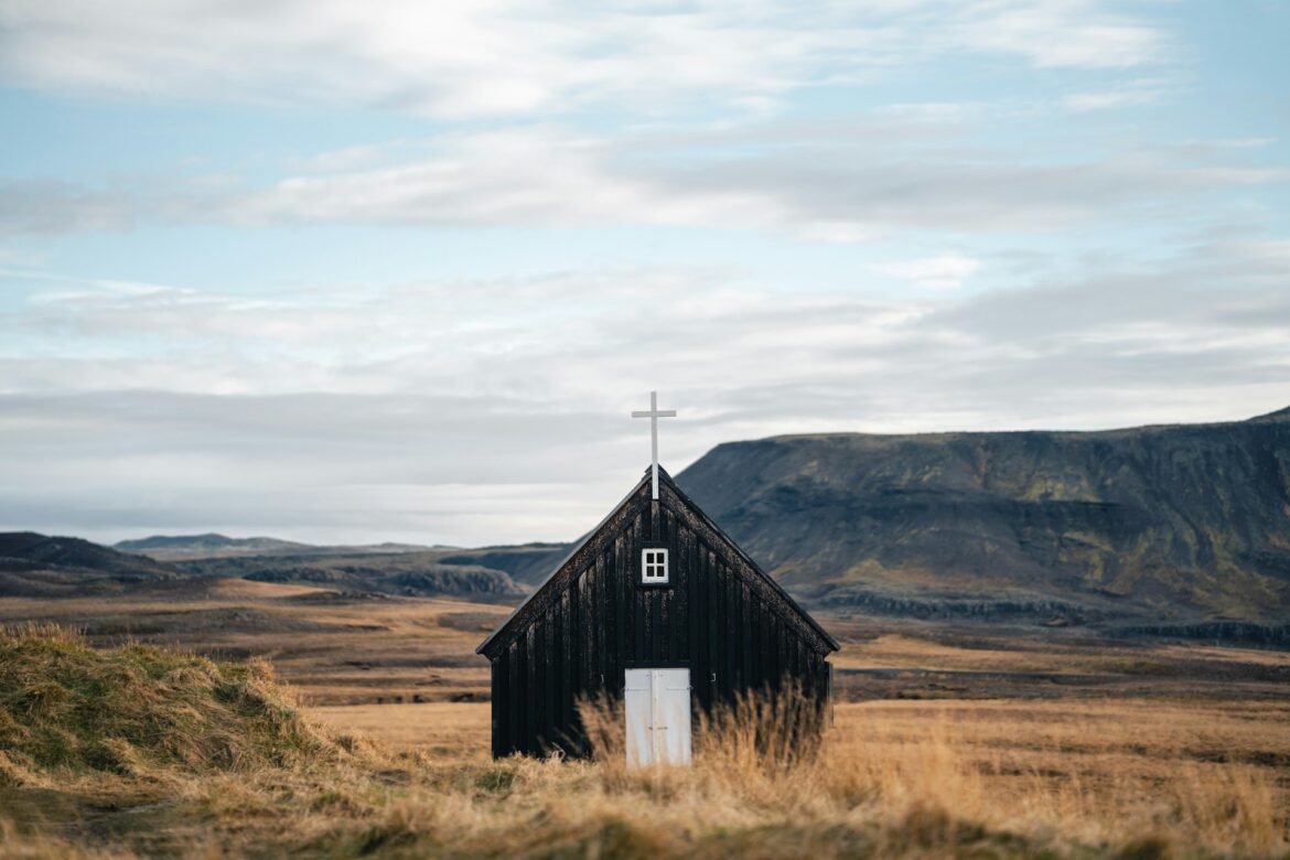 Black wooden chapel with a white cross on its peak, set in a windswept, open moorland with rolling hills in the background.