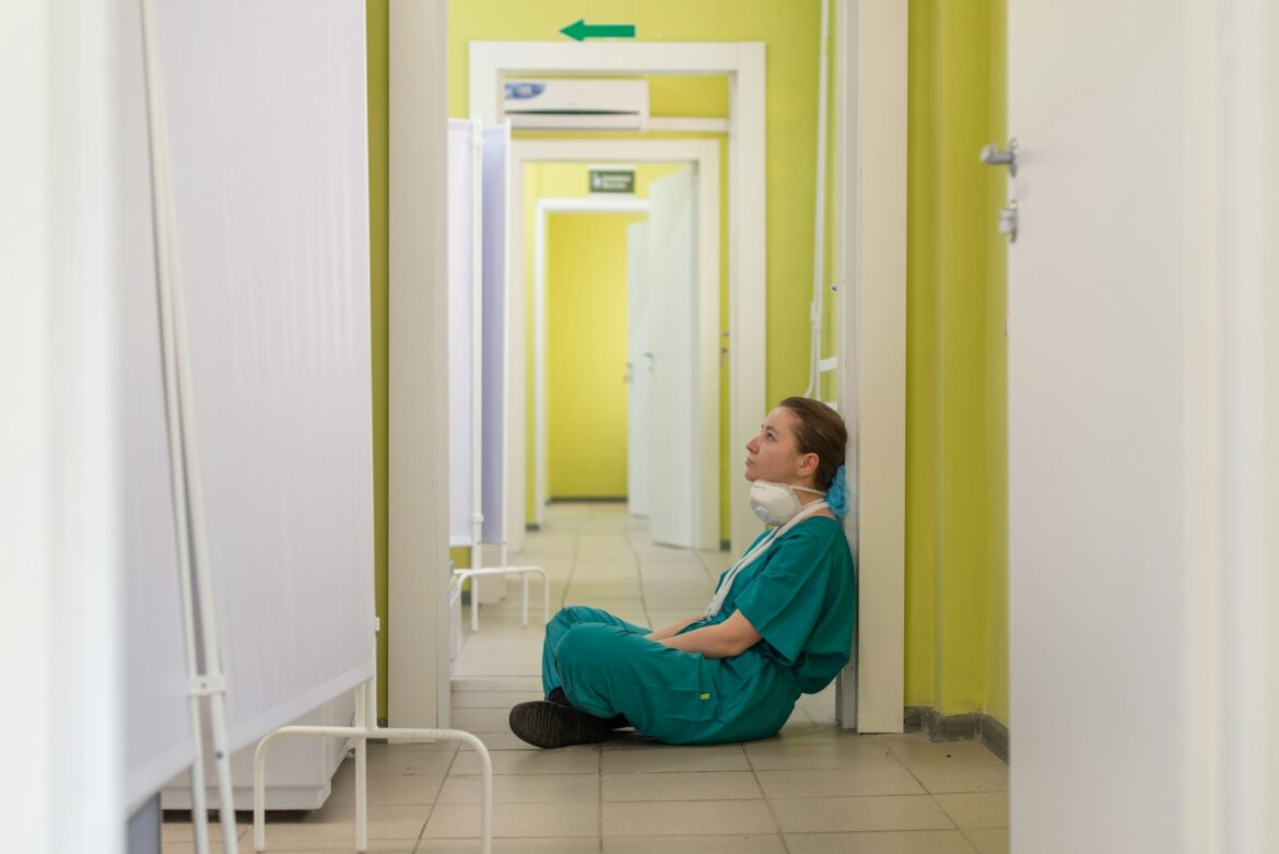 Healthcare worker in teal scrubs sits against a bright yellow hospital hallway, wearing a mask and hair covering.