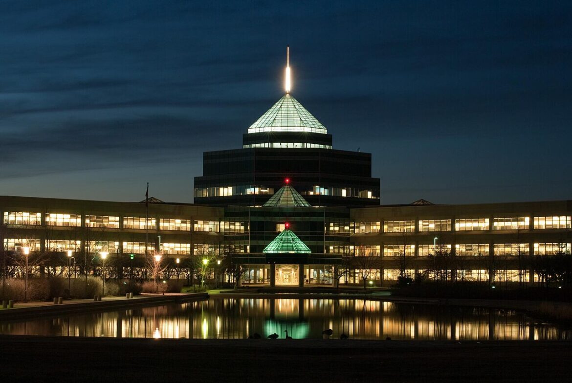 Night view of a modern, glass-roofed building complex lit from inside, with a central glass pyramid and reflection in a foreground pool.