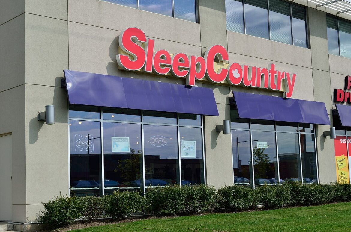 Sleep Country storefront with a large red logo, purple awnings, and glass windows with shrubs in front.