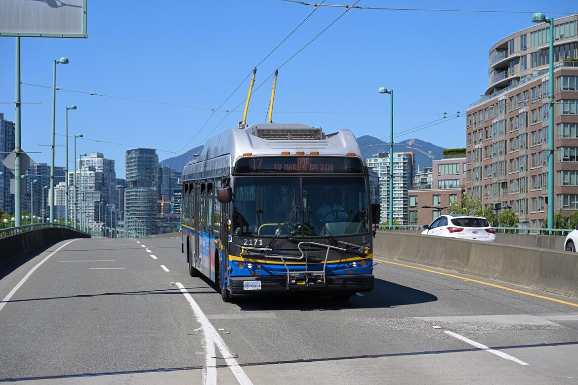 Silver and blue trolleybus driving on a city bridge with overhead power lines and a skyline of tall buildings behind it.