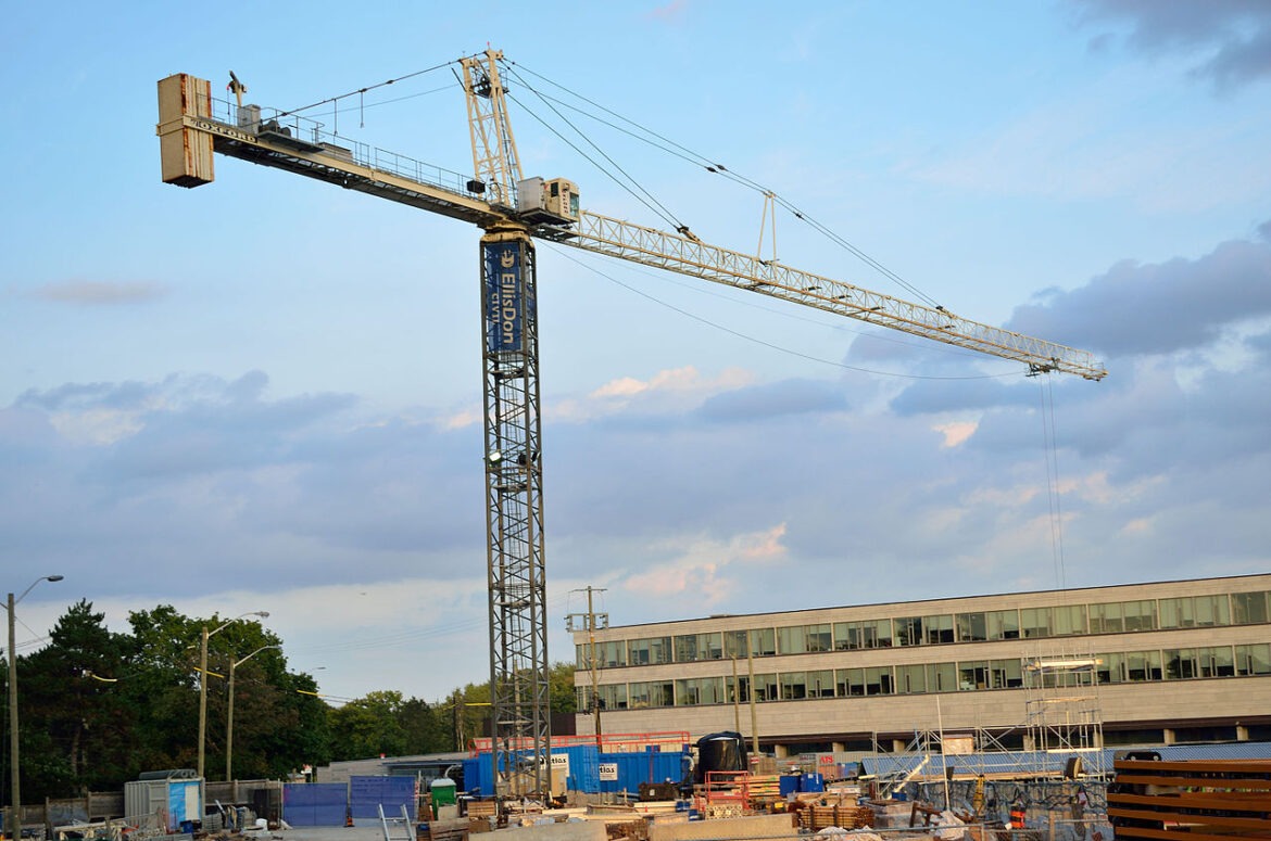 Tower crane over a construction site with a mid-rise building in the background and scattered materials and equipment on the ground.