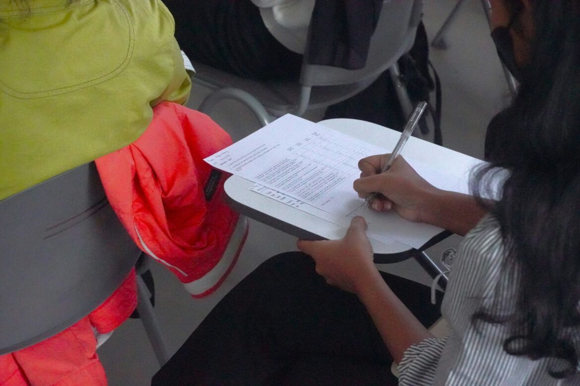Person signs a form on a clipboard resting on their lap, with a bright hi-vis jacket nearby.