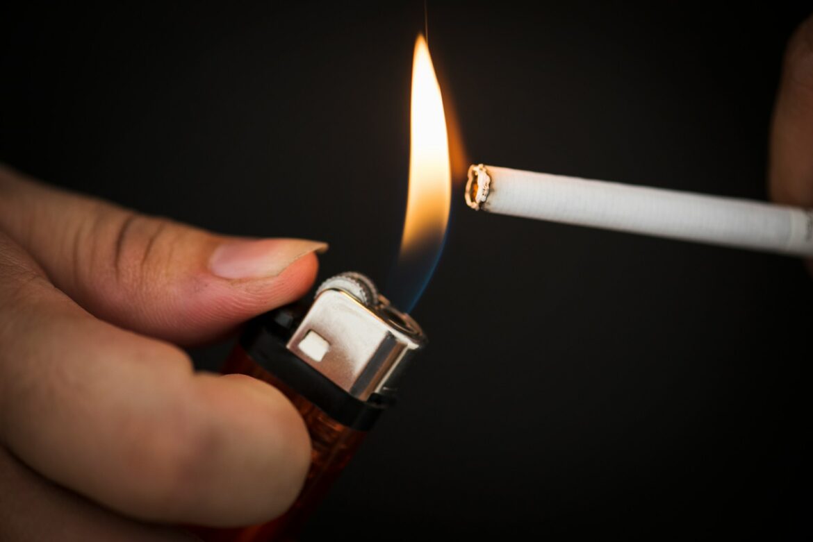 Close-up of a hand lighting a cigarette with a lighter, flame to the end against a dark background.
