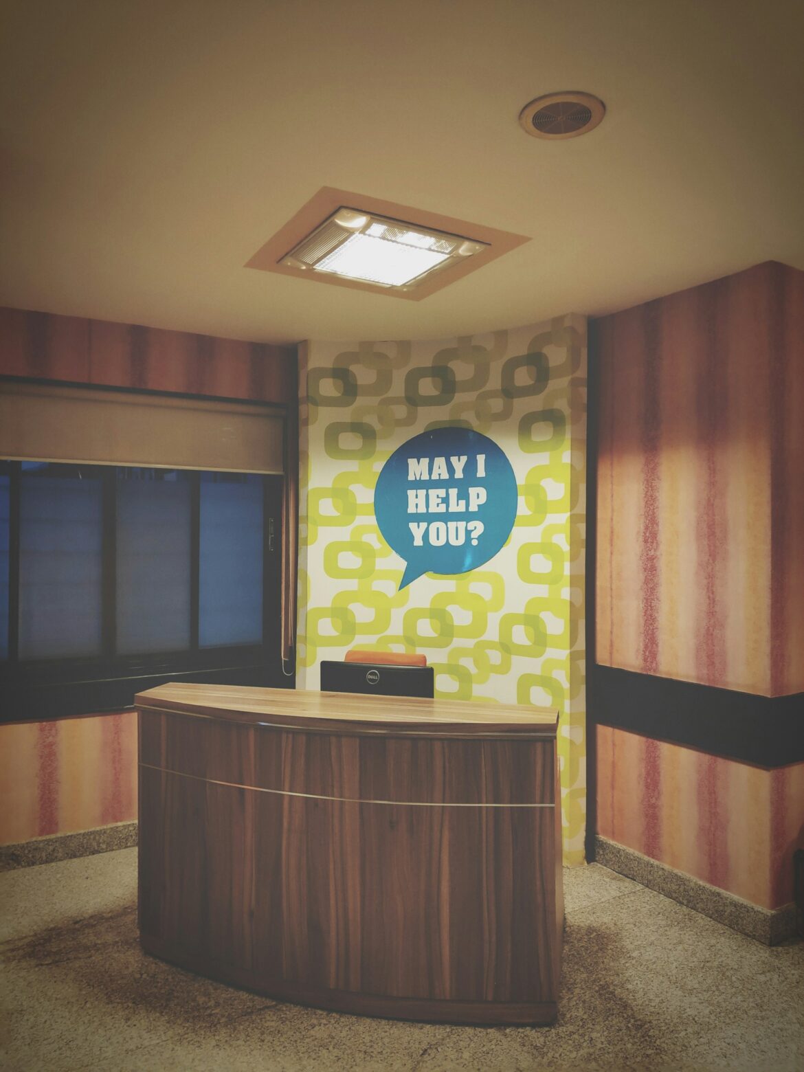 Reception desk in a warm lobby with a patterned wall and a blue speech-bubble sign reading 'MAY I HELP YOU?'