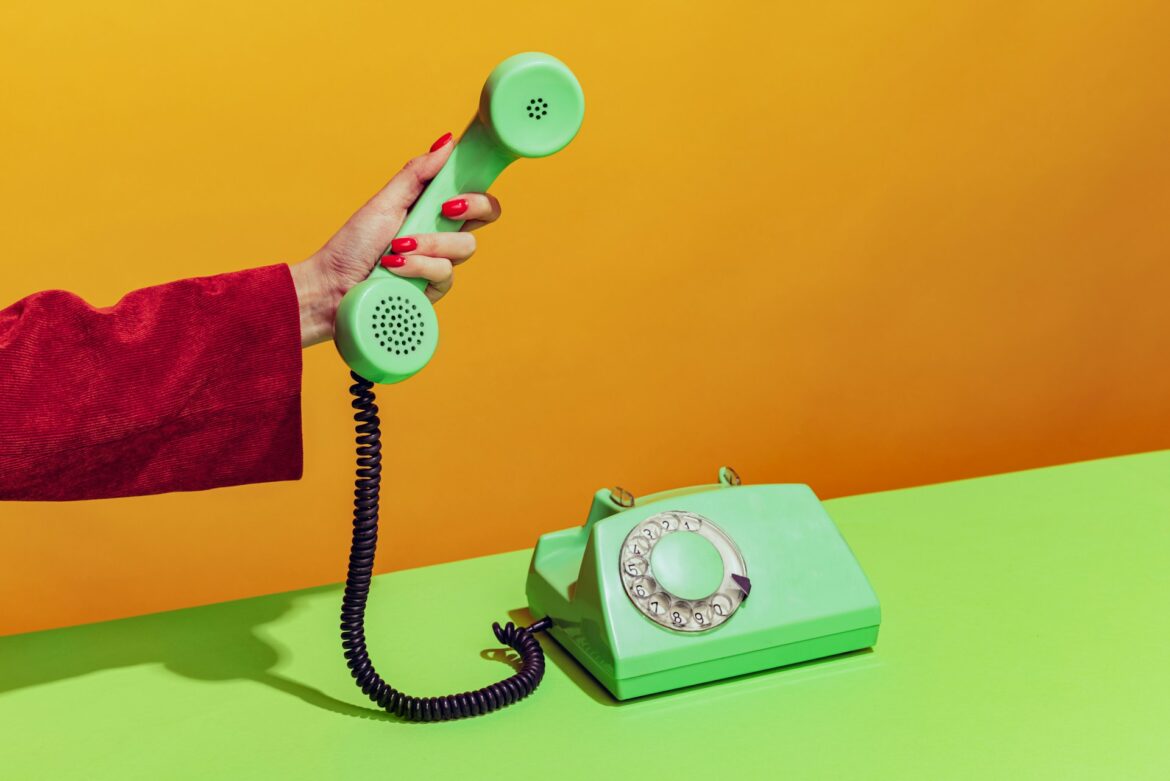 Hand holding a green telephone receiver over a lime-green desk against an orange wall, in red-sleeved sleeve.