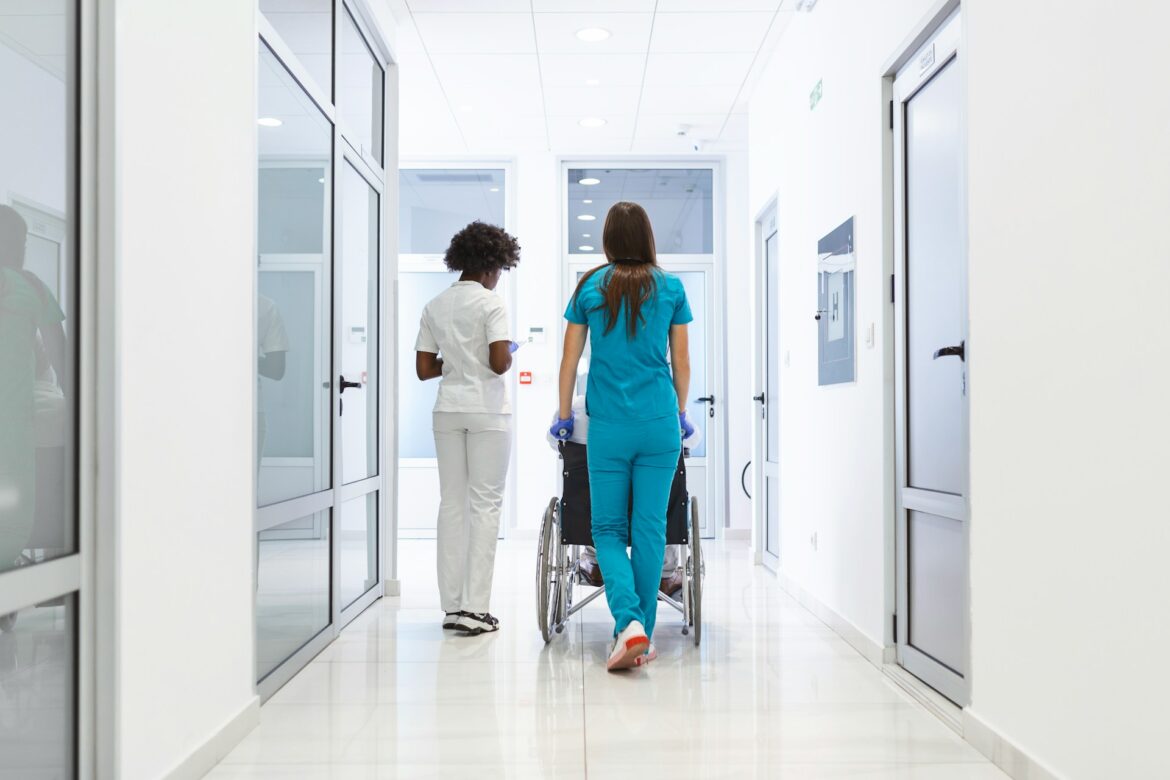 Two medical workers push a patient in a wheelchair down a bright hospital hallway.